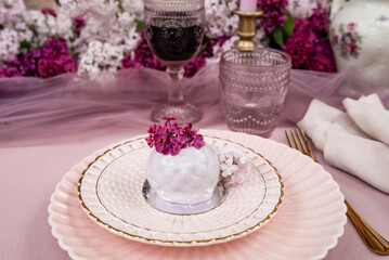 Fototapeta premium Elegant glazed dessert decorated with lilac blossoms on a pastel pink table setting. Romantic spring celebration concept with flowers, fine tableware and vintage atmosphere.
