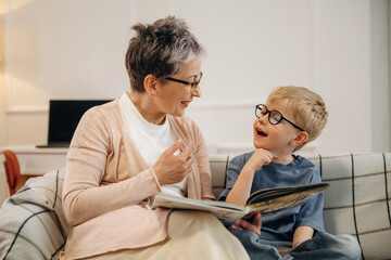 Fototapeta premium Elderly woman with short gray hair and glasses reads a book to a young boy with blonde hair and glasses while sitting on a couch in a cozy living room
