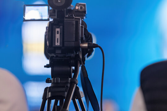 Professional broadcast video camera mounted on a tripod at an indoor media event, with a blue illuminated background and blurred figures suggesting an active live production session.