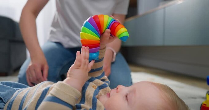Adorable baby lying on the floor playing joyfully with a colorful toy while a caregiver watches closely in a cozy home environment, capturing moments of early childhood development and happiness