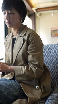Soft light streams through train window. Woman sits calmly, eyes downcast, fingers resting gently. Coat is tan, lapels soft, button gleaming. Patterned seat beside her adds quiet texture
