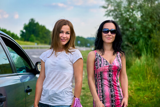 Two young cheerful women near car on road during trip