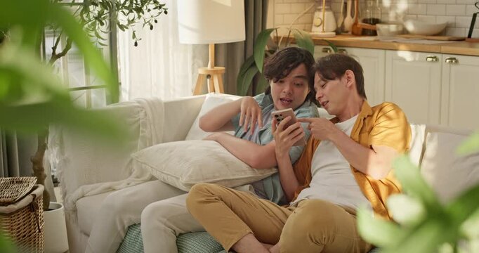 A playful young Asian gay couple relaxing on a couch, smiling and looking at a smartphone screen sharing online content or checking social media at home. LGBTQ+ relationship, diversity lifestyle.