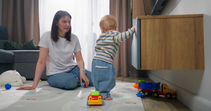 A young mother engaging with her toddler son during playtime on a carpeted floor at home, encouraging exploration and interaction with toys and household objects in a bright living room setting