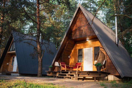 A-frame log chalet house in the forest with a natural backdrop. Summer getaway in a wooden cottage. Outdoor terrace, seating area, firewood