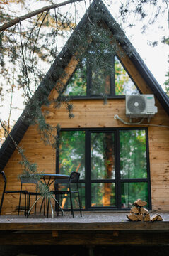 A-frame log chalet house in the forest with a natural backdrop. Summer getaway in a wooden cottage. Outdoor terrace, seating area, firewood