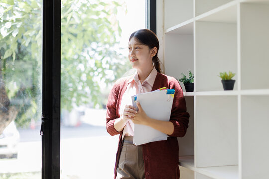 Young asian professional woman holding documents. Standing by a window. Looking ahead with a thoughtful and optimistic expression