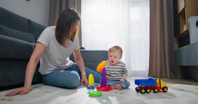 A mother and her baby playing together on the floor with colorful stacking rings and toy trucks, engaging in early childhood development activities in a cozy living room setting filled with natural 