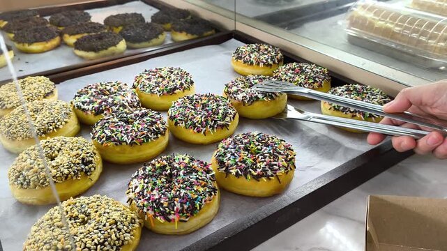 A customer picks up sweet donuts from a display case in a bakery using serving tongs.