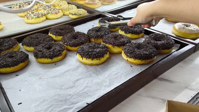 A customer picks up sweet donuts from a display case in a bakery using serving tongs.