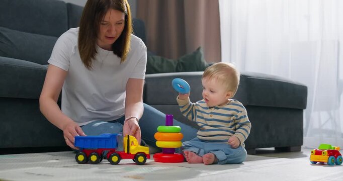 Mother and baby playing together on the floor with colorful stacking rings and toy trucks in a cozy living room, promoting early childhood development and bonding