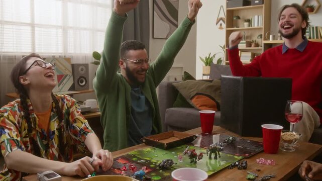 Young biracial man in glasses throwing dice and cheering while playing RPG tabletop game with group of friends in cozy living room