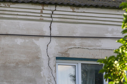 Large deep vertical crack on the old concrete stucco facade of a residential house wall running down from the roof line, building structural failure and property insurance concept.