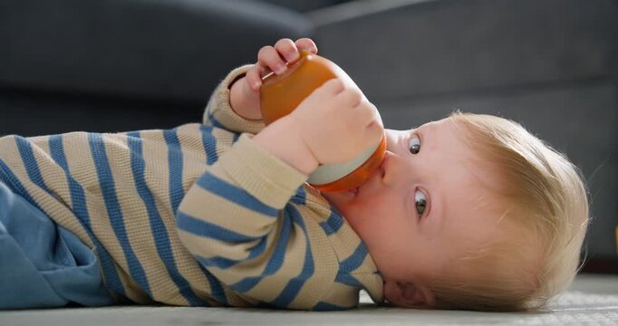 A baby boy lying on the floor drinking from a sippy cup while wearing a striped long-sleeve shirt and blue pants, captured in two frames showing initial and final moments of drinking.