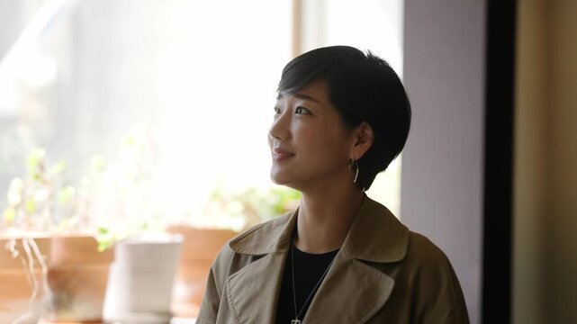 Woman smiles, touching a potted plant. Soft sunlight illuminates her gentle expression. Window frames cozy indoor gardening scene. Her calm gaze suggests quiet enjoyment. Ideal for tranquil, home