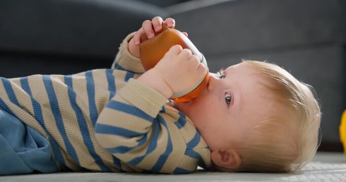 Adorable infant lying on the floor wearing a striped sweater and blue pants while drinking from an orange sippy cup, capturing a tender moment of early childhood hydration and comfort