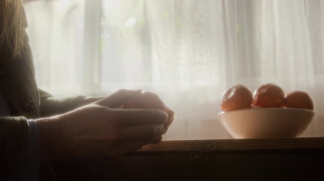 Side view close up shot of unrecognizable young woman peeling tangerines at kitchen table against white window curtains, cozy slow living scene lit with hazy lighting, copy space