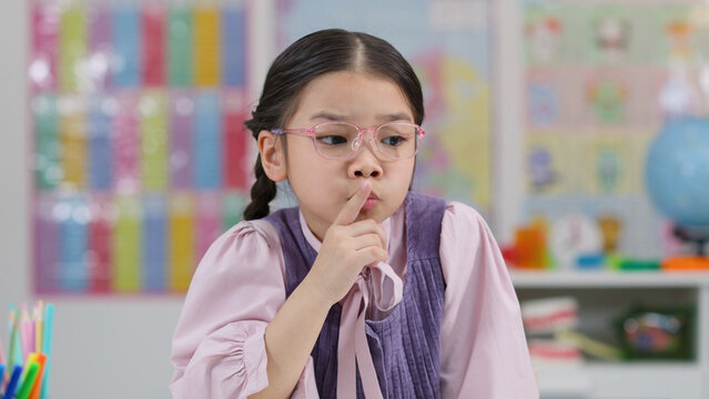 Young East Asian Schoolgirl with Glasses Making Quiet Shh Gesture in Classroom