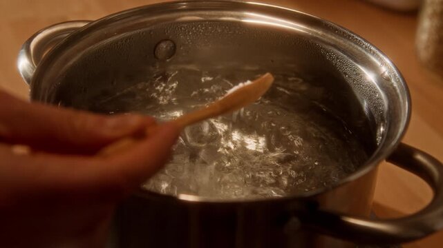 Close up shot of unrecognizable young woman dropping teaspoon of granulated salt into bubbling boiling water while cooking on gas stove in tiny home kitchen, copy space