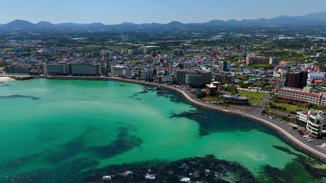 Aerial flyover of Hamdeok bay sweeping over a turquoise lagoon, traditional stone fish traps and a seaside town toward the volcanic coast, Jeju Island, South Korea
