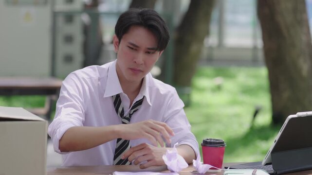 A serious-looking Asian man is checking his resume and digital tablet while searching for a new job at an outdoor table in a park.