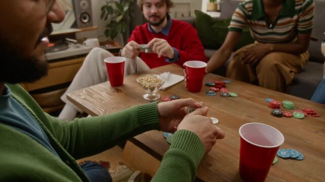 Tilt up shot of young biracial man shuffling cards, sitting on floor at wooden coffee table, enjoying evening with friends, playing games