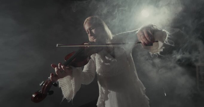 Video frame of a violinist in a white sweater playing in smoky studio light, with soft backlighting, dark background, atmospheric haze, and copy space