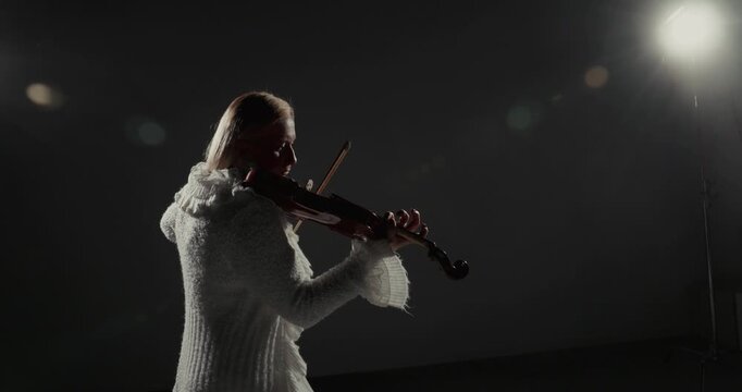 Video frame showing a violinist in white performing under a bright studio light against a dark low-key background with copy space