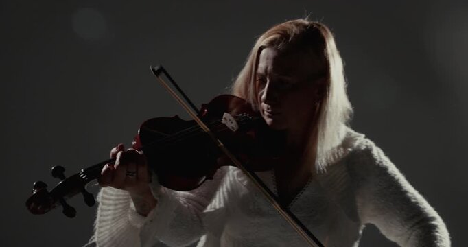 Video frame showing a violinist in white performing under a bright studio light against a dark low-key background with copy space