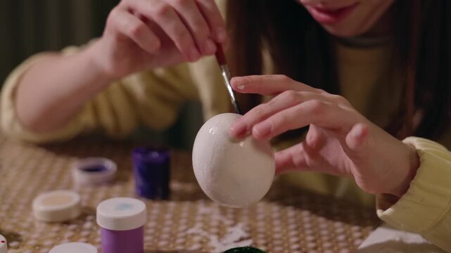 Young woman painting decorative patterns on white easter egg with purple gouache at home.
