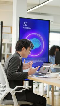 Asian businessman or programmer working on a computer while looking at AI assistant interface on monitor at office. Smart workplace automation, future software engineering, machine learning concept.