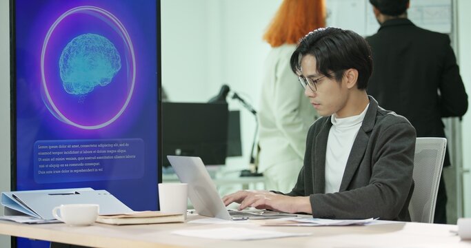 Asian businessman or programmer working on a computer while looking at AI assistant interface on monitor at office. Smart workplace automation, future software engineering, machine learning concept.