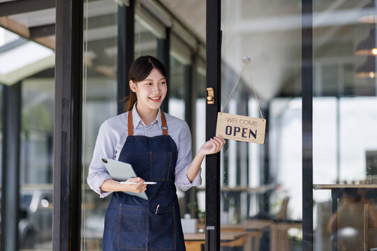 Woman entrepreneur standing at store entrance, smiling and flipping welcome open sign, inviting customers to new small business cafe or restaurant startup