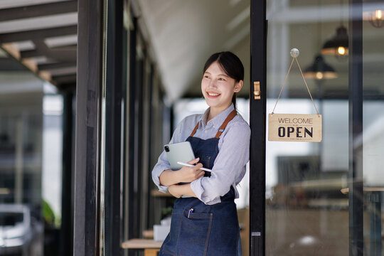 Young smiling entrepreneur standing at the entrance of a cafe, holding a tablet and a digital pen, ready to welcome customers, representing small business ownership and success