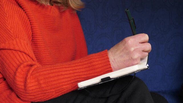 Close-up of woman hands writing a grocery list in a notebook. The scene shows everyday planning and domestic organization.