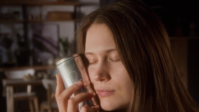Close up shot of young woman holding drink in aluminum can with no label and pressing cold metal surface to skin refreshing in summer heat indoors and enjoying sensation, copy space