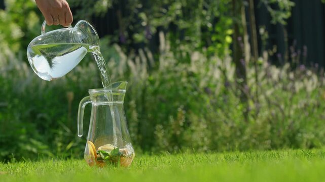 Clear water pouring from glass pitcher into a larger pitcher with sliced citrus fruits and mint leaves on green grass in sunlit garden.