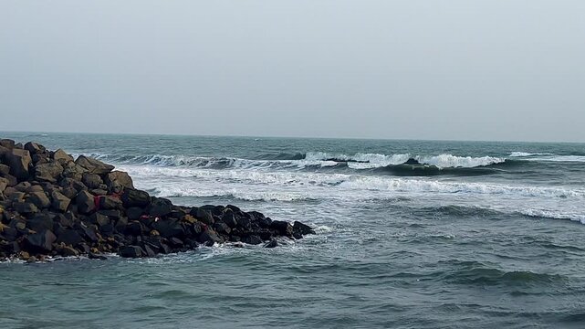 Wide-angle view of Arabian Sea swells breaking along dark rocky jetty at Dwarka. Ideal for coastal India travel, pilgrimage docs, ocean power, and moody atmospheric seascape content.