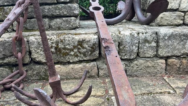 A close-up shot pans across several large, rusty ship anchors and chains displayed outdoors against a weathered stone wall