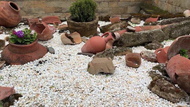 A collection of broken terracotta pots, clay fragments, and ancient artifacts are scattered on white gravel, with a potted plant in the foreground