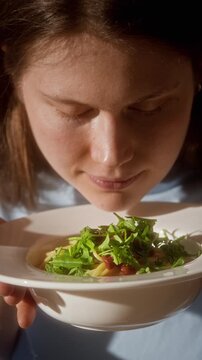 Vertical front view shot of smiling young woman smelling freshly cooked homemade pasta dish enjoying simple meal in sunlight, story format