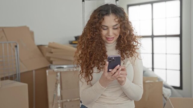 Woman holding smartphone and smiling while surrounded by cardboard boxes and ladder in a building; contentment moving day.