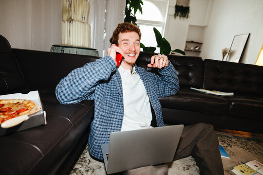 The young adult man moves between laptop and smartphone tasks in a relaxed living room.