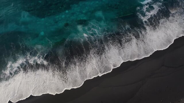 Turquoise wave crashing onto smooth black sand beach with foamy surf aerial view under soft sunlight serene coastal scene