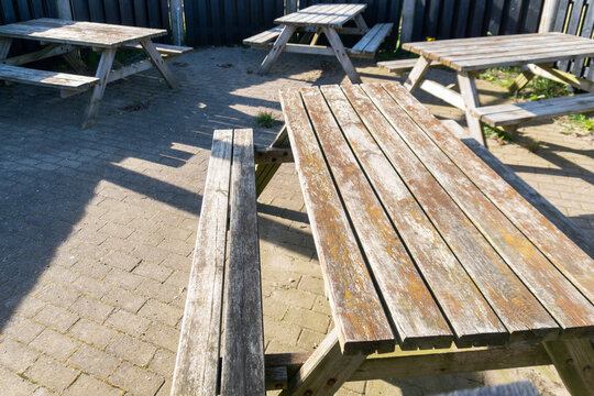 Weathered wooden picnic tables and benches stand on paved ground in a sunny outdoor seating area, showing worn boards, shadows, and rustic public recreation furniture.