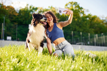 Fototapeta premium Young woman with curly hair takes selfie with her border collie dog while sitting on green grass in a sunny park with trees in the background
