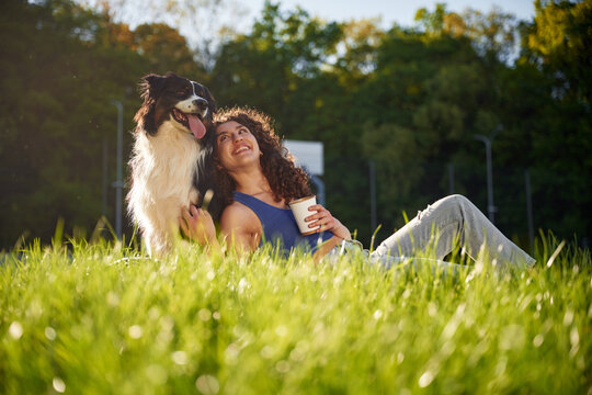 Young woman with curly hair sits in green grass, enjoying a sunny day outdoors with her happy dog, holding a cup and smiling at the pet beside her