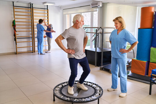 Senior Man On Trampoline With Physiotherapist Supervising In Rehab Center For Injury Recovery And Balance Training Indoors
