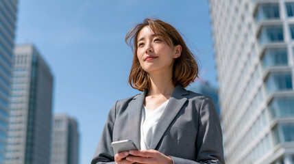 Fototapeta premium Professional Woman with Phone Downtown: A focused professional woman in a grey suit stands outdoors amidst modern city buildings.