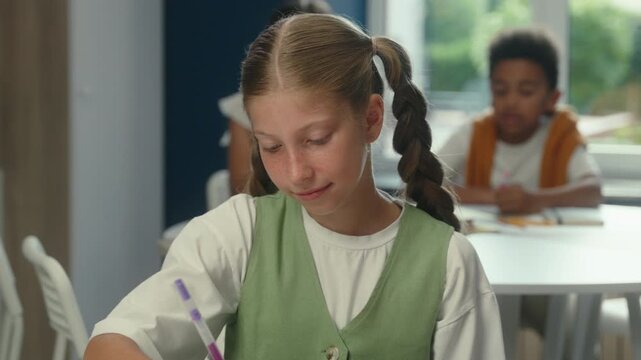 Caucasian happy little girl kid child schoolgirl pupil writing in copybook at class lesson course educate studying learning at desk write classwork in classroom near multiethnic pupils classmates